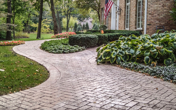 Curved brick-pattern stamped concrete walkway bordered by planting beds beside a brick house